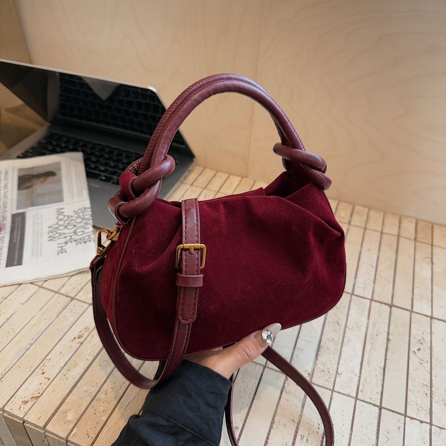 Burgundy handbag held by a person with a laptop and magazine in the background.