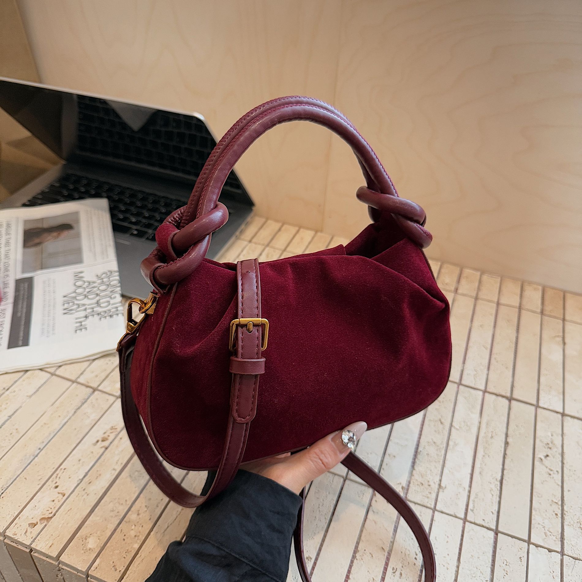 Burgundy handbag held by a person with a laptop and magazine in the background.